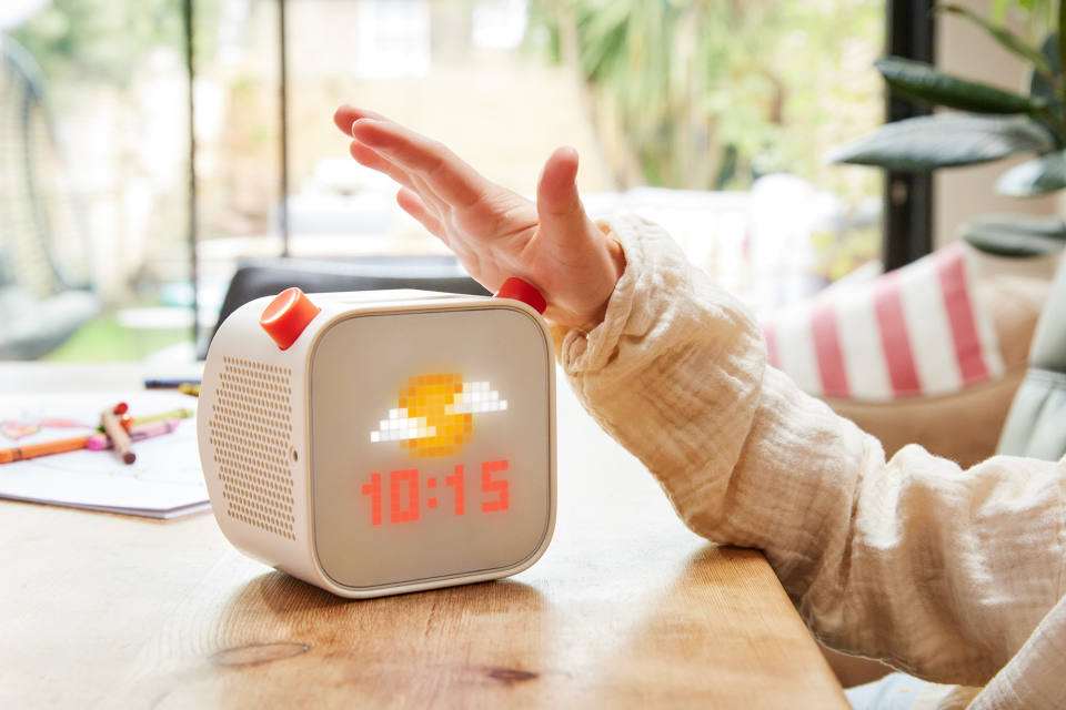 A person presses a button on a Yoto Player, which is displaying the time and a pixellated image of a sun that's partially obscured by clouds.