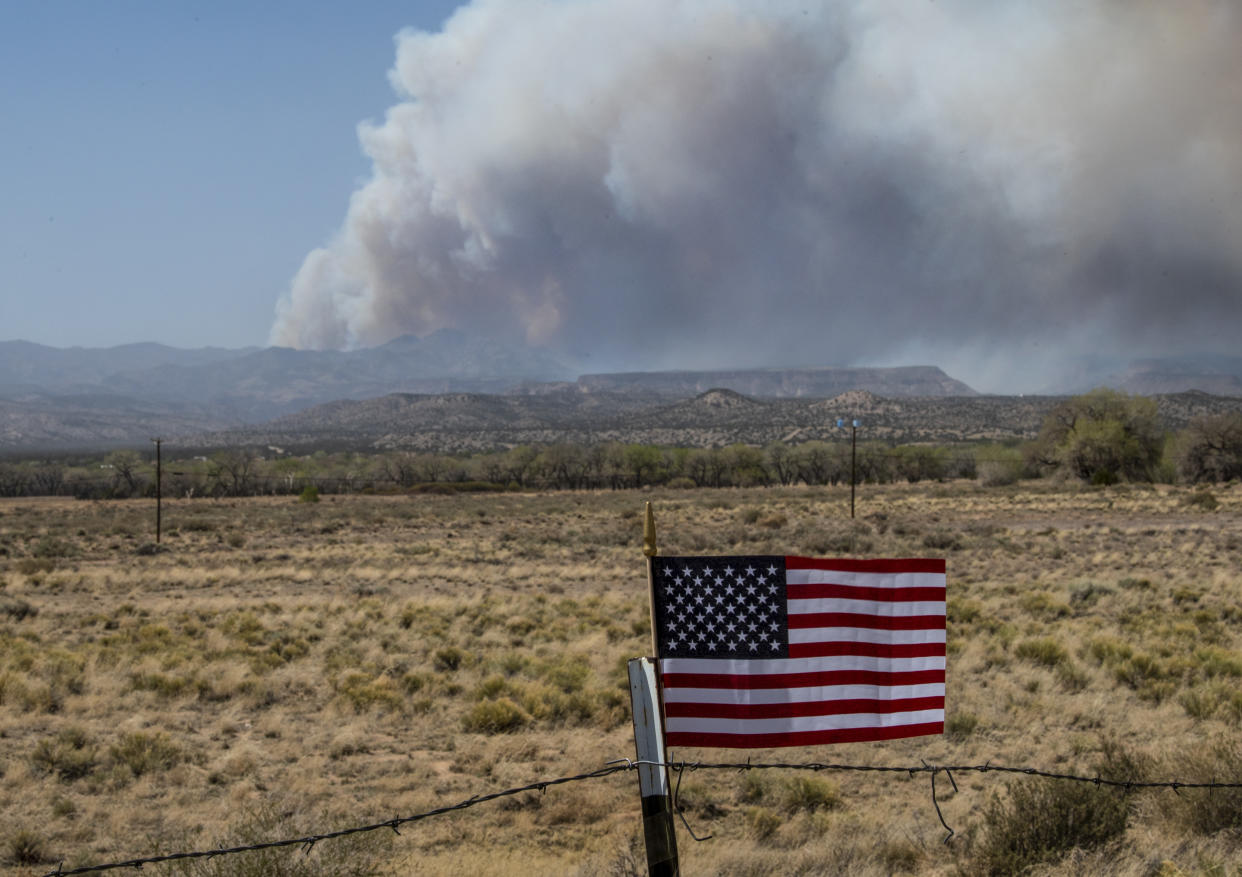 An American flag on a fence blows in the wind as heavy plumes of smoke billow in the distance.