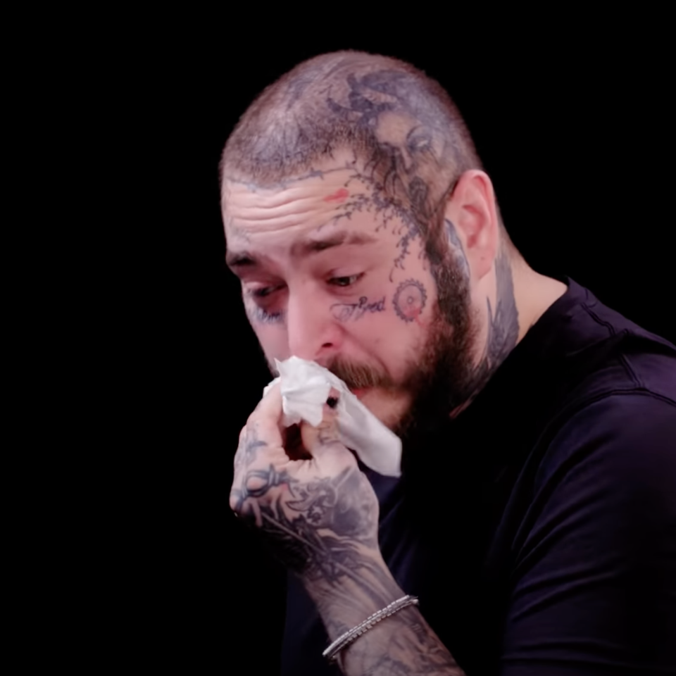Post Malone wipes his nose with a tissue while appearing emotional on a black background