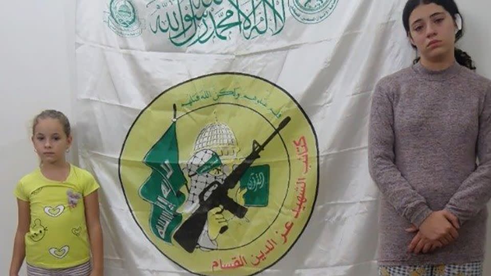 The IDF released an image showing sisters Ela and Dafna Elyakim, who were previously held hostage by Hamas, in front of a flag with the Hamas logo. - IDF