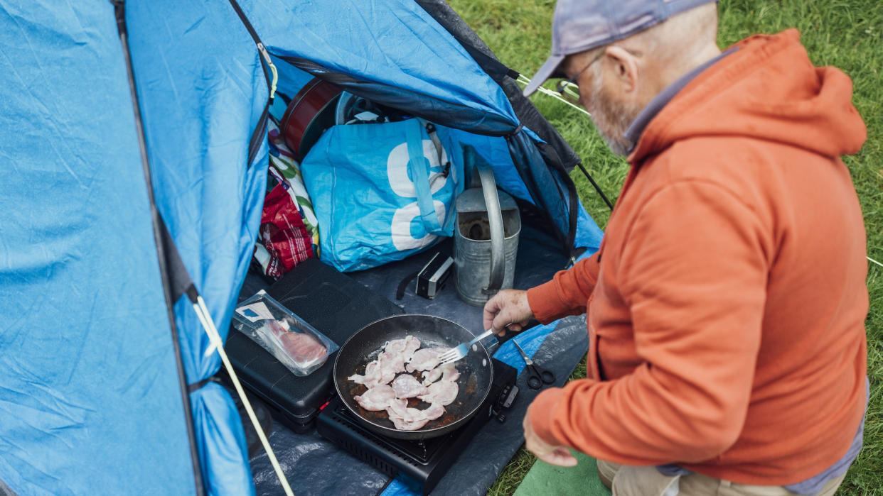  An over the shoulder view of a senior man kneeling down by his tent to cook bacon in a portable camping stove, he is preparing breakfast at his campsite at Hollows Farm, The Lake District in Cumbria, England. 