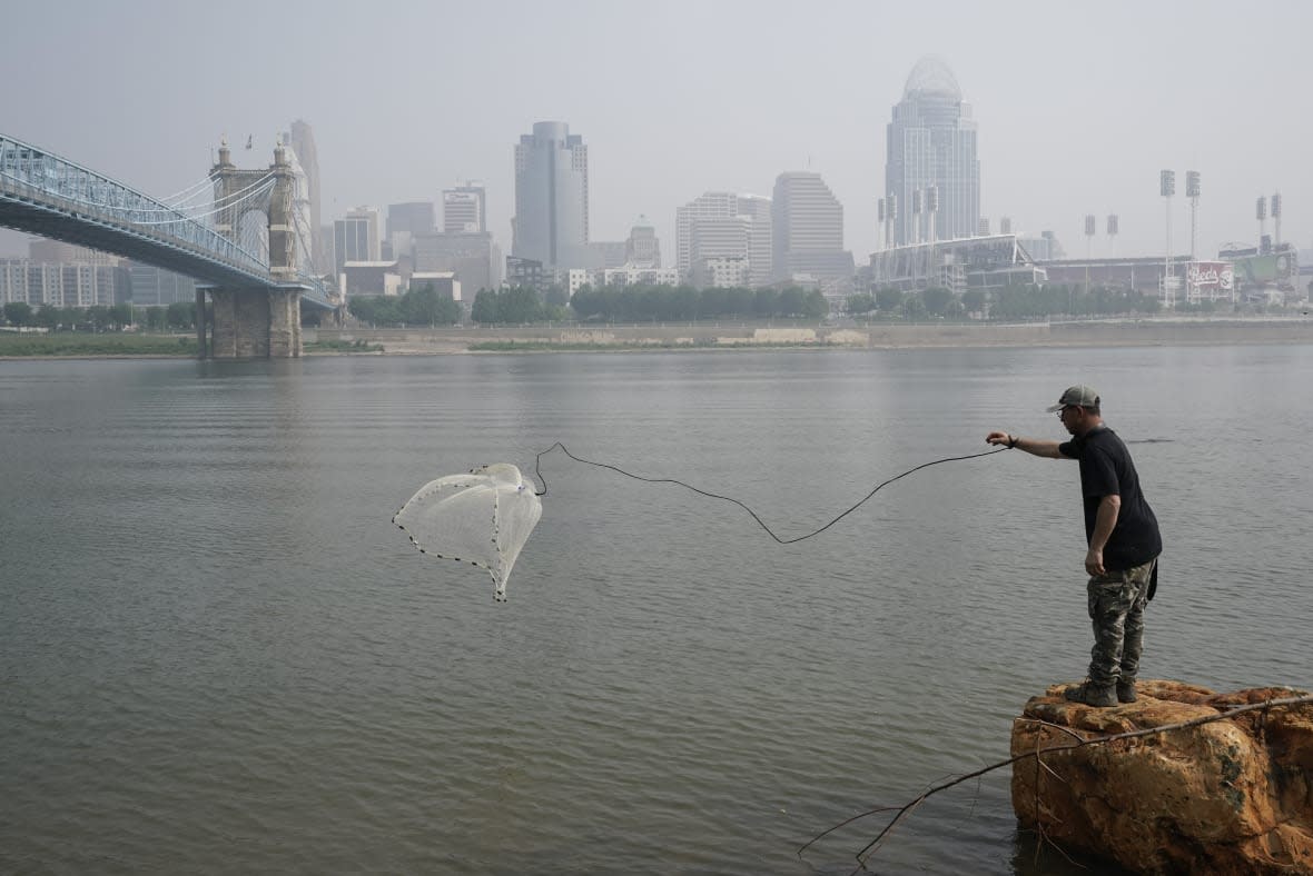 A man tosses a fishing net into the Ohio River as smoke from wildfires is visible over downtown Cincinnati, Wednesday, June 28, 2023. (AP Photo/Joshua A. Bickel)