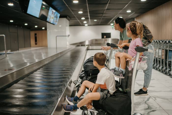 A family of four, including two children, waits at an airport baggage claim area. One child sits on a luggage cart, while the others watch the conveyor belt