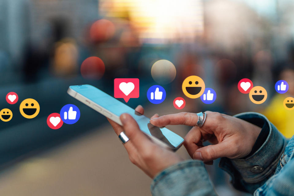 Close-up of hands holding a phone with floating emojis, indicating social media interaction related to love and approval