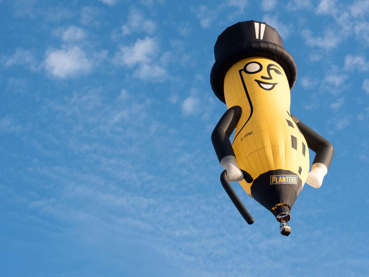 a hot air balloon shaped like Planters' Mr. Peanut Mascot, a yellow peanut with black arms, a black cane, a monocle, and a black top hat, soars in the middle of a blue sky