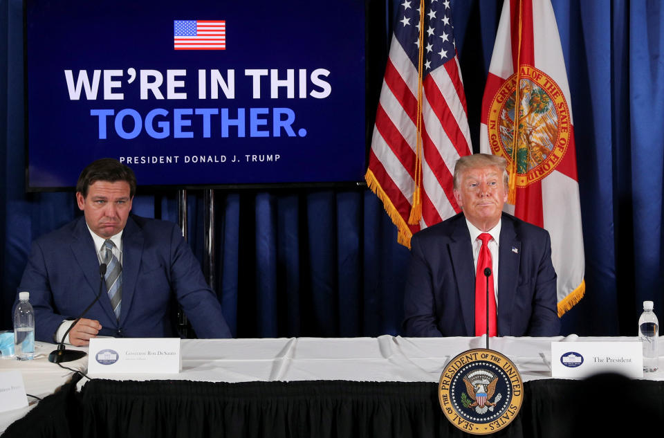 Gov. Ron DeSantis appears to scowl next to Former President Donald Trump at a table with microphones in front of various flags
