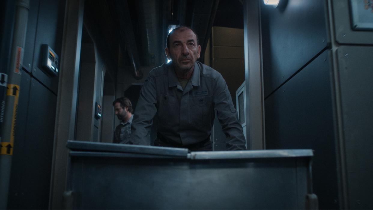  A man in gray work coveralls embellished with a 'Helios' logo is pushing a metal cart down an industrial-looking corridor. 