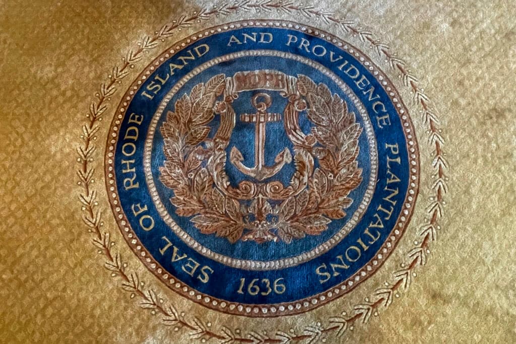 FILE – The state seal with Rhode Island’s full former name appears on a rug in the state room on Nov. 4, 2021, at the Rhode Island State House, in Providence, R.I. A new report issued Monday, Aug. 22, 2022, suggests ways Providence can atone for its extensive ties to the transatlantic slave trade and centuries of racism and discrimination by, among other things, establishing home repair funds, launching financial literacy programs and boosting aid to Black and Indigenous organizations. (AP Photo/Jennifer McDermott, File)