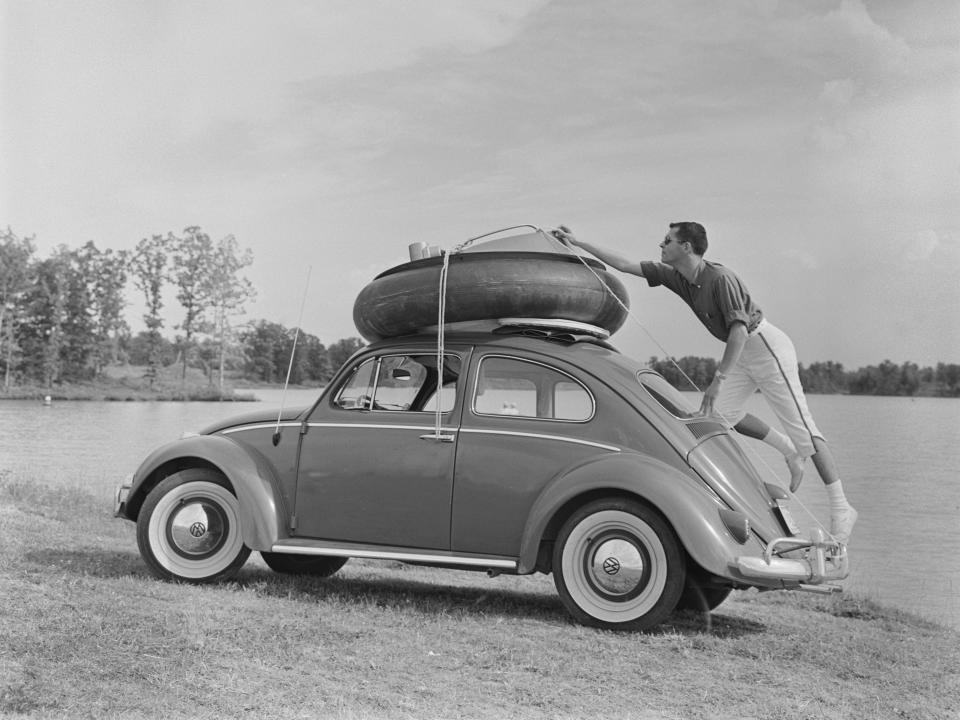 man stands on the rear bumper of a Volkswagen Beetle as he adjusts the ropes tying an inner tube and dinghy adaptor, to roof of the car, location unspecified, United States, circa 1955.