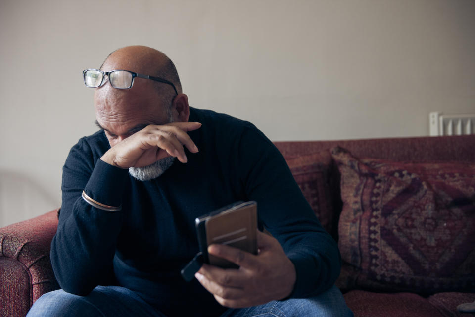 A man in a black sweater, seated on a couch, holds a smartphone in one hand and wipes tears from his eyes with the other hand