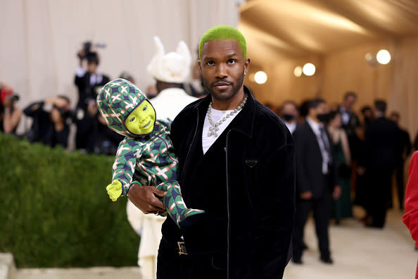 Frank Ocean at an event holding a green baby doll, with a neon green dyed buzz cut and a black velvet suit