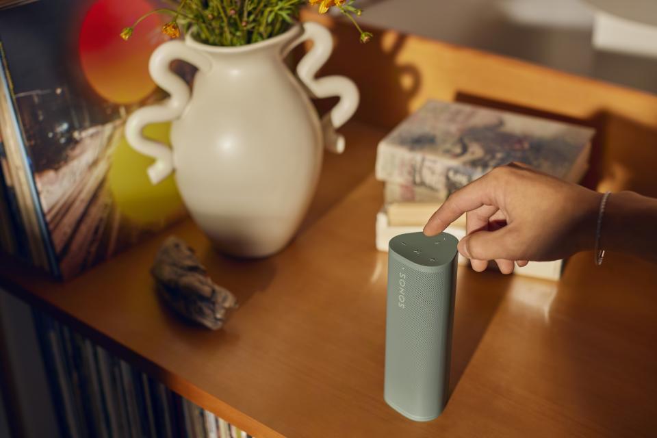 Fotografía de estilo de vida del altavoz portátil Sonos Roam 2.  Se encuentra en una mesita de noche ocupada mientras una mano alcanza el marco (desde la derecha) para tocar sus controles.
