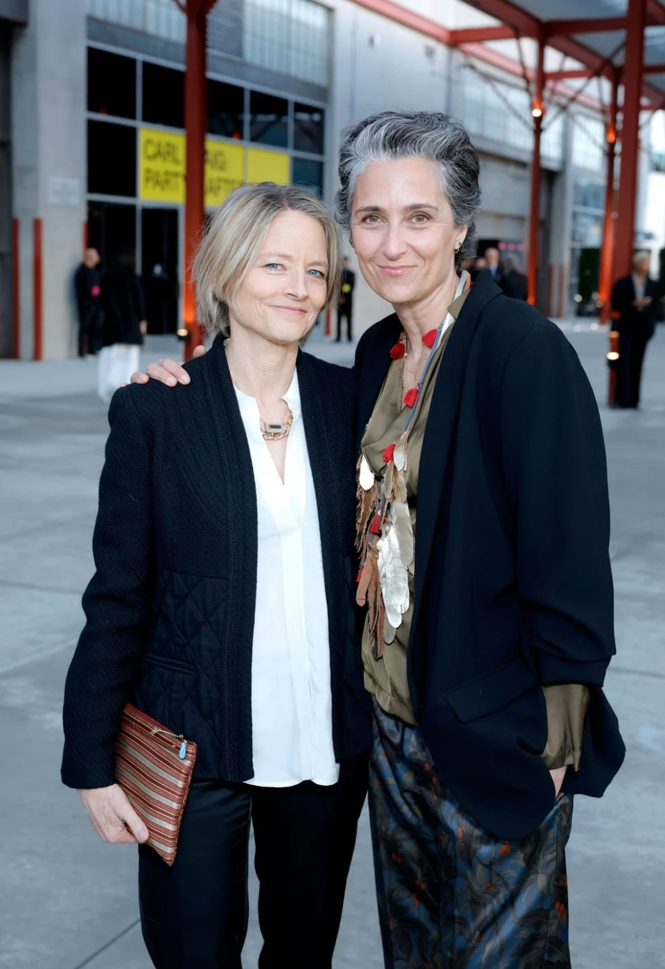 jodie foster and alexandra hedison stand next to each other, smiling and hugging for a photo, they bothe wear dark jackets over blouses