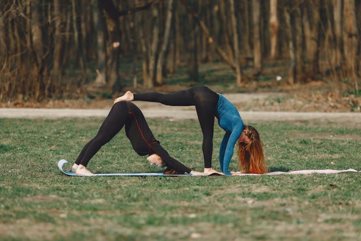 A woman in dark clothes folds forward in Downward Facing Dog pose while her yoga partner in a blue shirt does a variation of a standing split. They are practicing outside on grass.