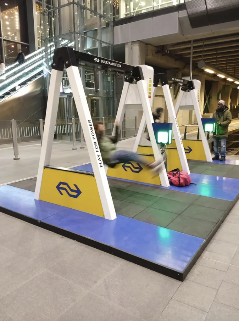 A person is swinging on an indoor public swing set with a logo that reads "NS" at a train station