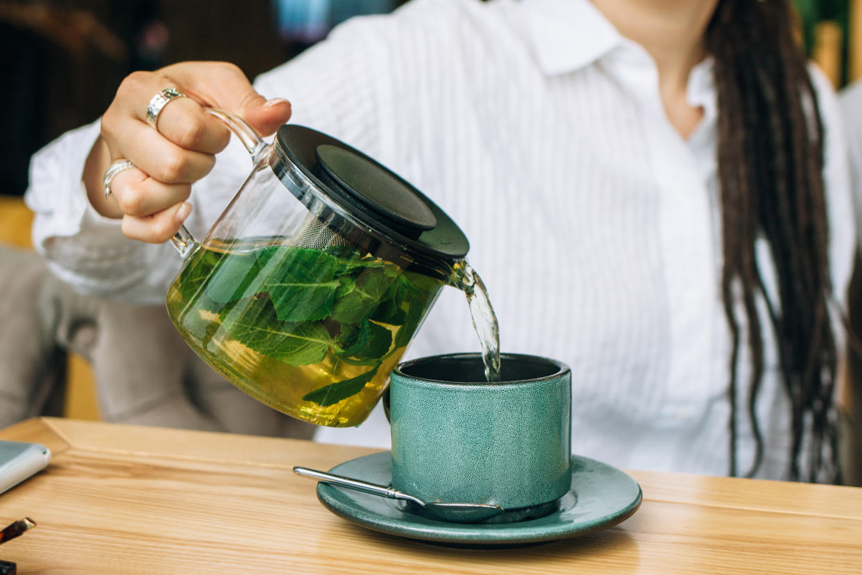 A woman pours green tea from a glass tea kettle into a green mug