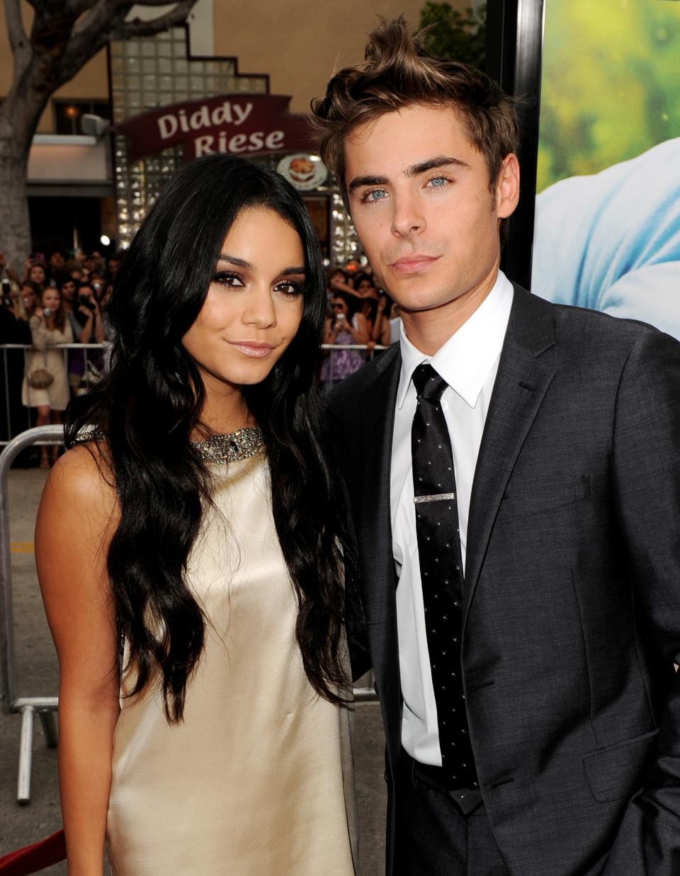 Vanessa Hudgens in a sleeveless dress and Zac Efron in a suit with a black tie pose together at an event