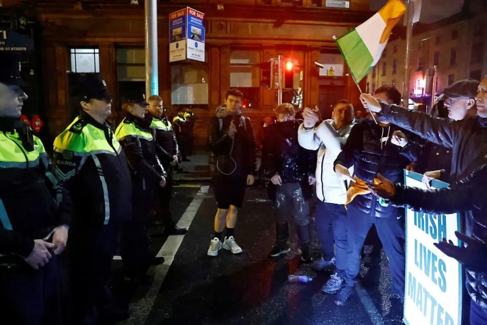 Varias personas permanecen de pie frente a la policía antidisturbios, cerca del lugar de un presunto apuñalamiento que dejó varios niños heridos en Dublín, Irlanda. (Crédito: Clodagh Kilcoyne/Reuters)