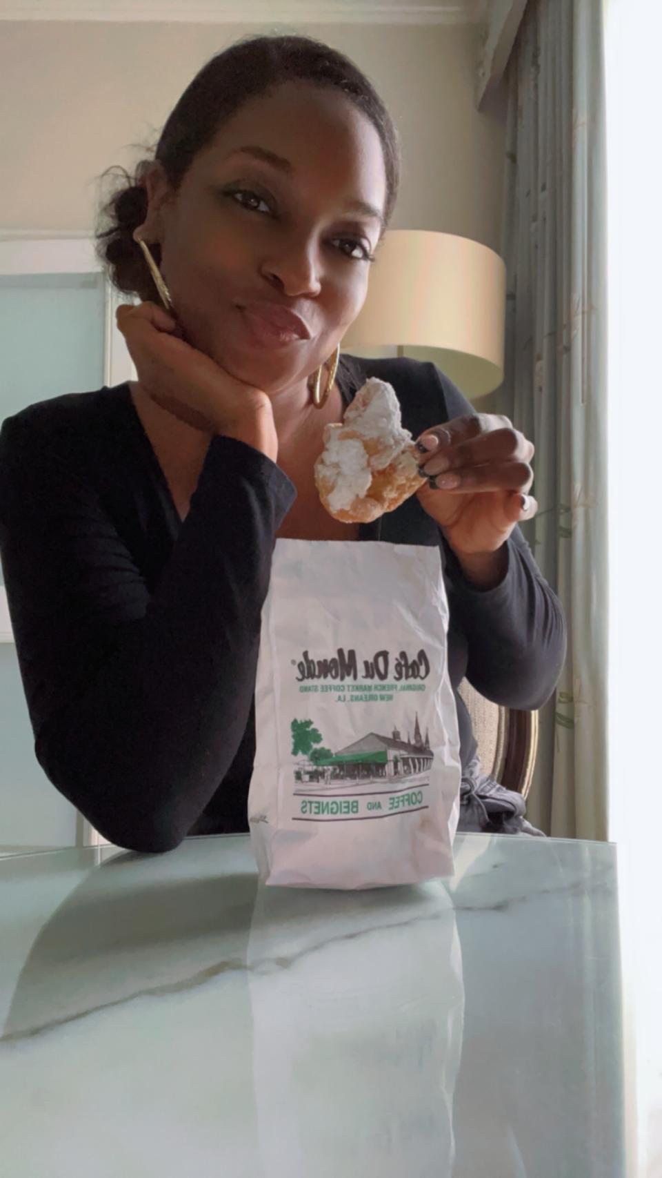 Woman holding beignet and leaning on table with Café Du Monde paper bag in front of her in a bright room