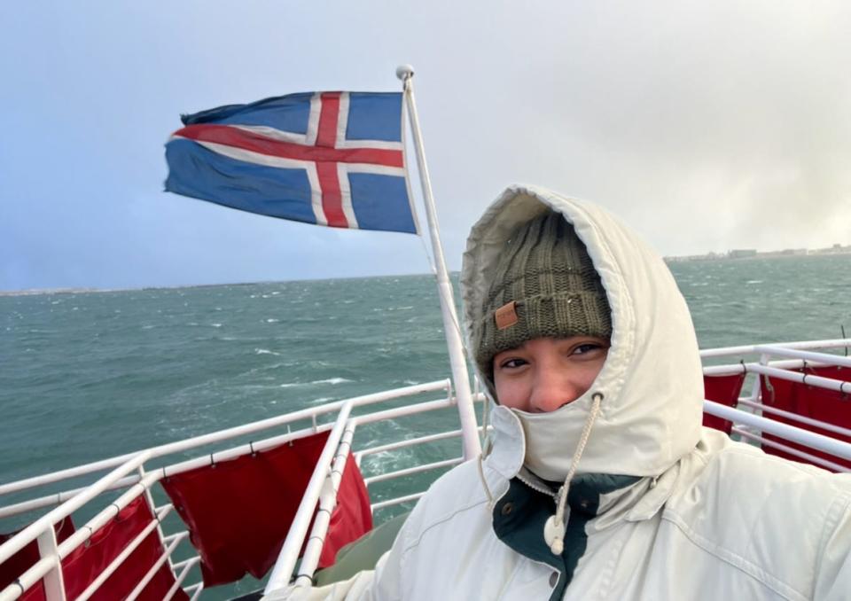 Selfie of the writer, wearing a white coat and a gray hat, on a ship with a white railing and a flag of Iceland