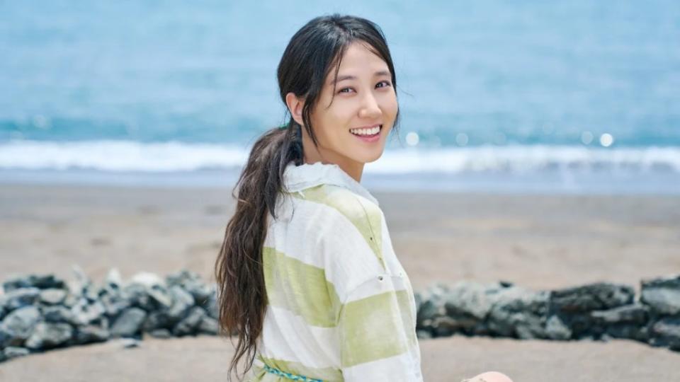 A woman sits on the beach in a striped shirt, her back to camera, looking over her right shoulder with a smille