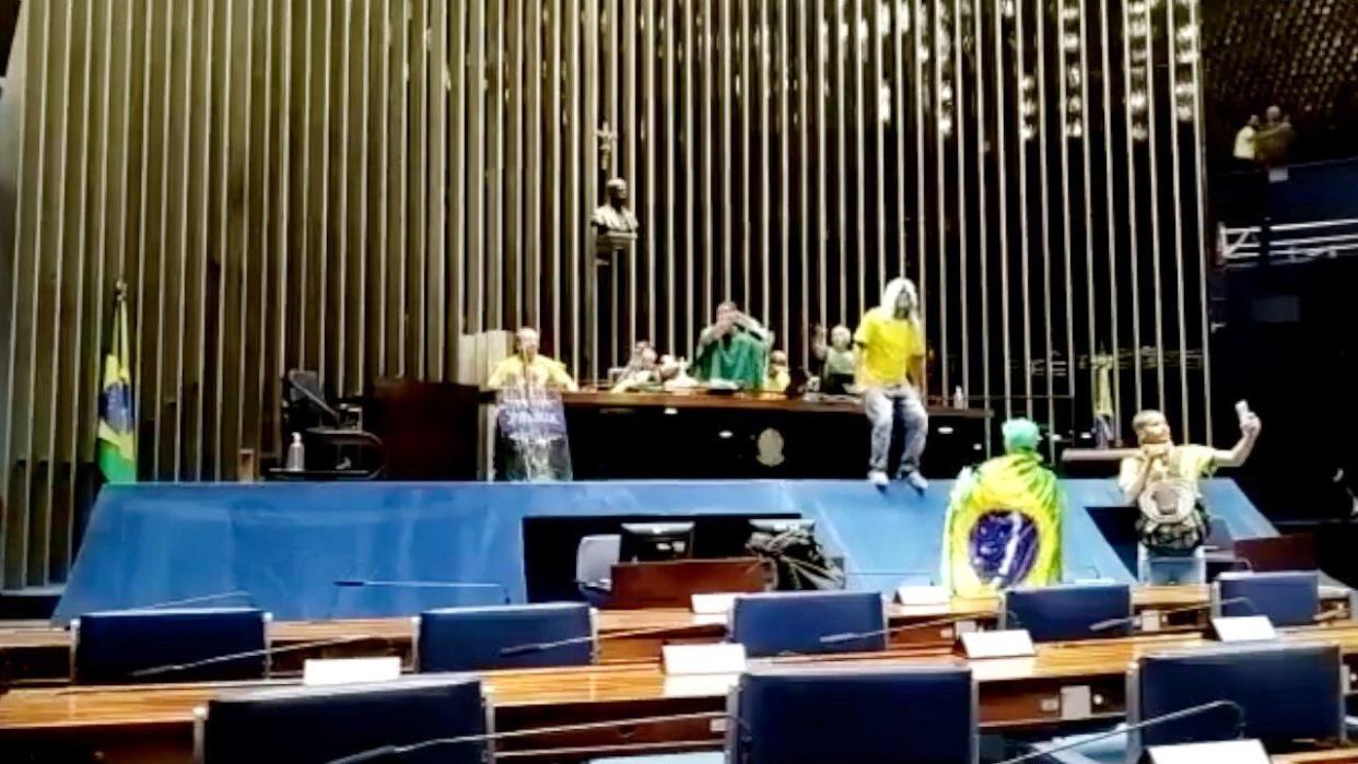 Supporters of former president Jair Bolsonaro rampage through the Federal Senate. One protester sits on a central desk with his feet on a protective barrier in front of it, another is draped in a Brazilian flag, and another films themself and the others with a cellphone..