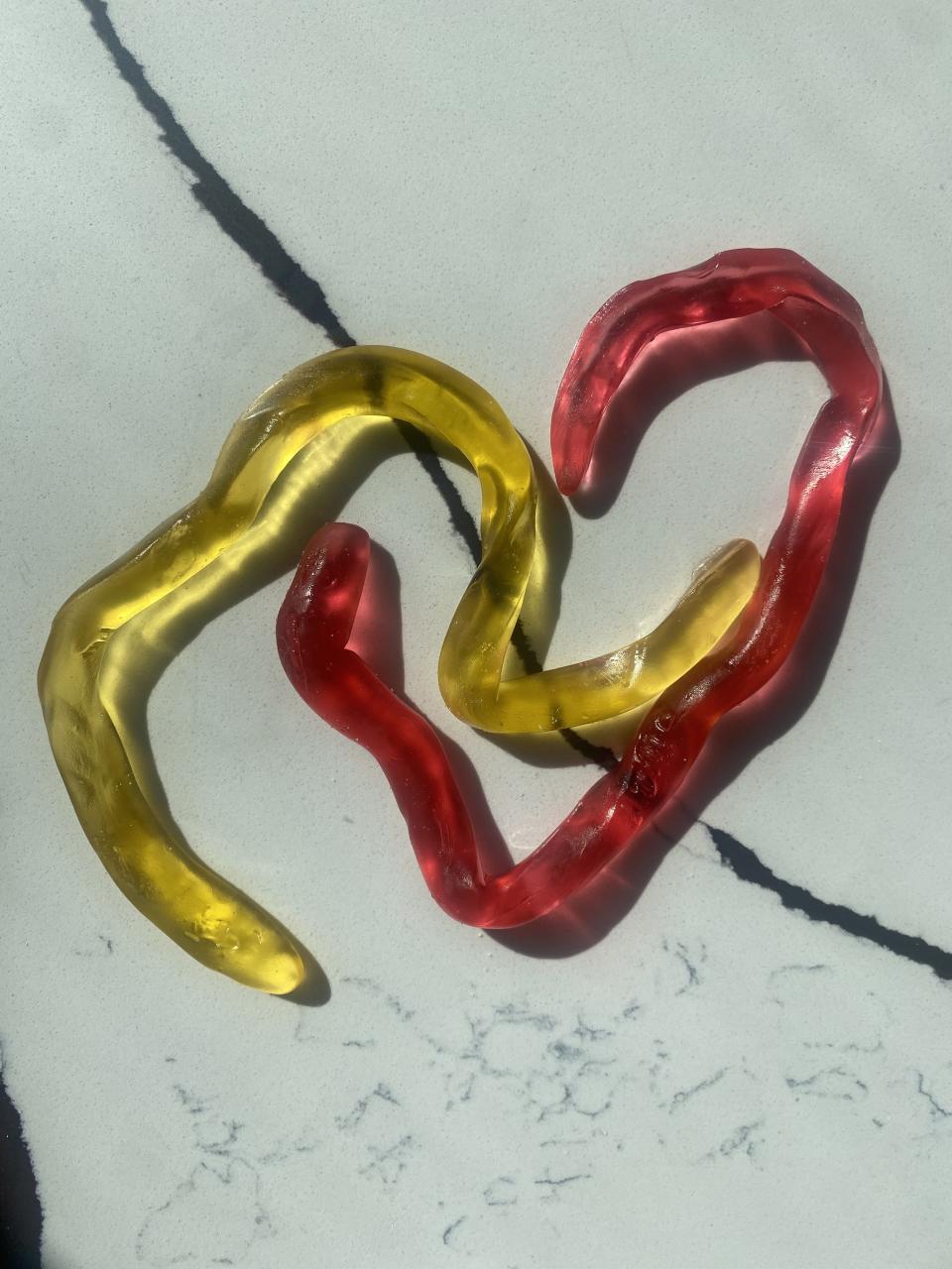 Two gummy snakes, one yellow and one red, intertwined on a marble surface forming a heart shape