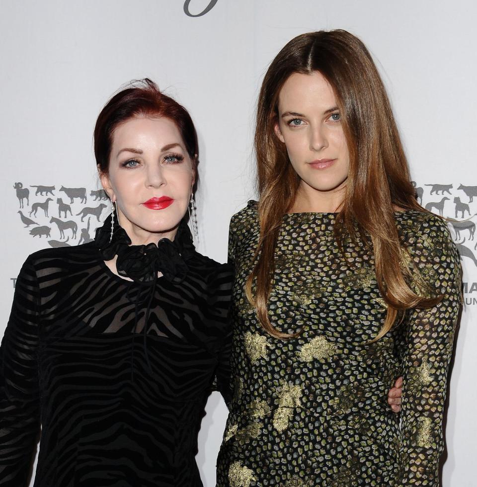 priscilla presley and riley keough pose for a photo together and smile at the camera, presley wears a black transparent dress, keough wears a green and black patterned long sleeve dress