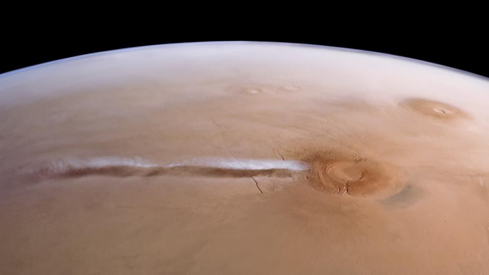  Wispy clouds float above a reddish orange planet as seen from space. 