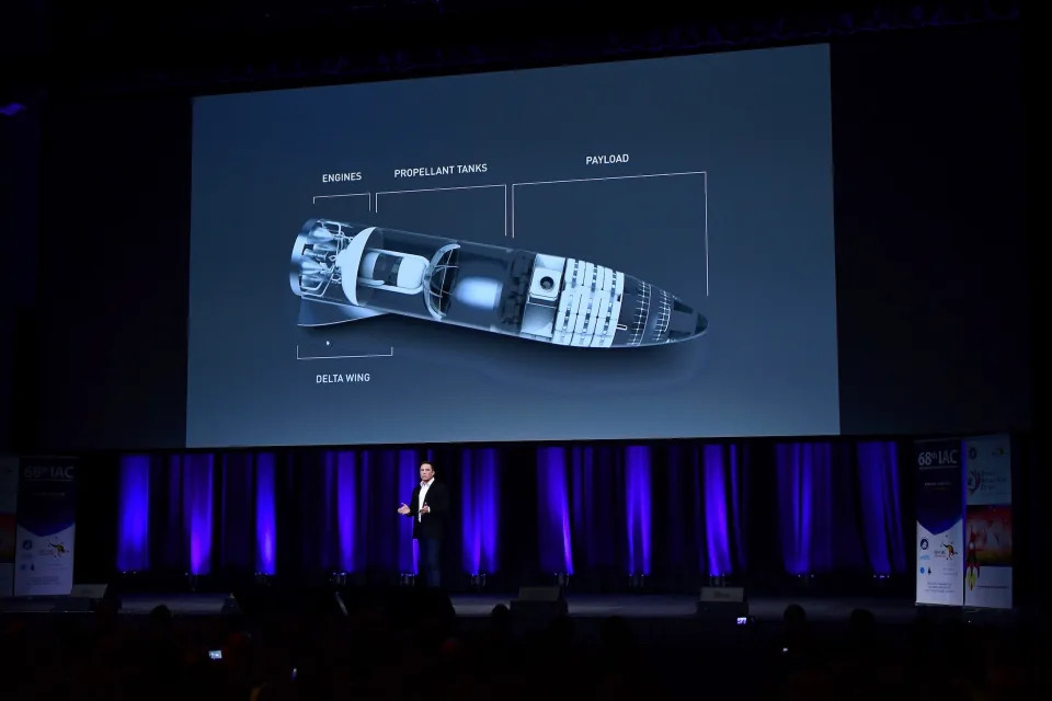 Elon Musk on a stage with dark blue curtains behind a large screen with an image of space travel vehicle labeled with different parts like engines, payload, and delta wings