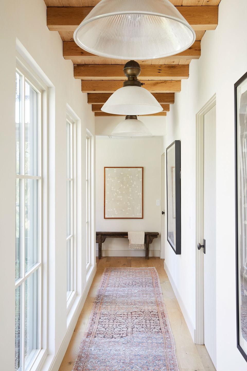 Four East-facing, floor-to-ceiling Marvin casement windows allow “the most magical morning light” to spotlight three oversize Obsolete Inc. Holophane glass and brass gas light pendants that hang next to Ross Alan reclaimed white oak lumber beams and above a multicolor vintage runner from Shoppe Amber Interiors.