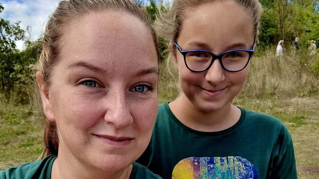 Close-up of a white mum and daughter's faces as they stand in a field. The daughter is wearing glasses with blue frames. Three figures stand in the distance under a tree.