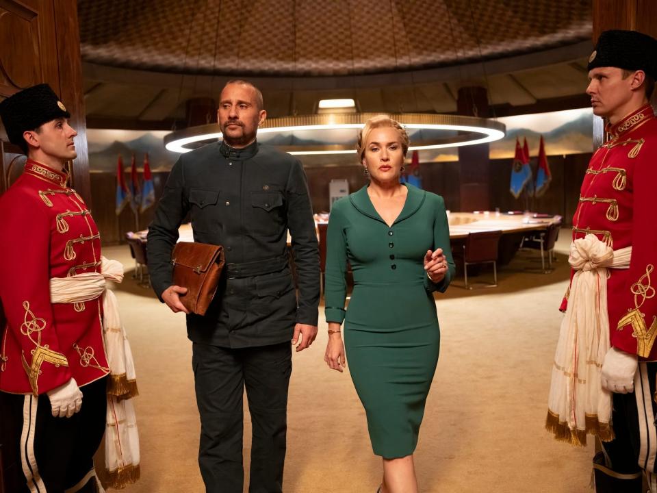 matthias schoenaerts and kat winslet in the regime, walking between two uniformed guards. schoenaerts is wearing military clothes, while winslet is in a form fitting, professional green dress