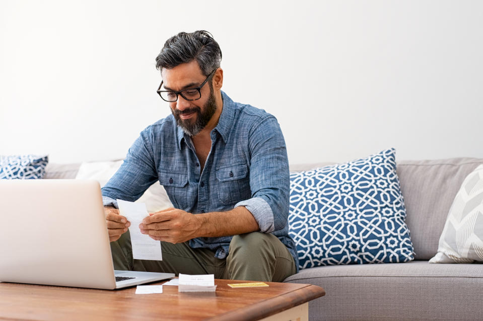 Mature casual man using laptop while looking at invoice. Smiling latin man managing finance with bills and laptop while sitting on couch at home. Hispanic guy reading expenses wearing eyeglasses.