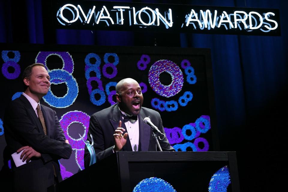 Michael Shepperd, alongside David Tarlow, speaks into a microphone on a stage with the sign "Ovation Awards."
