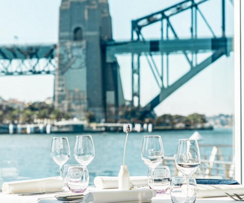 Glassware on a white restaurant table with sunny views out to the blue water with Sydney harbour bridge in the background.