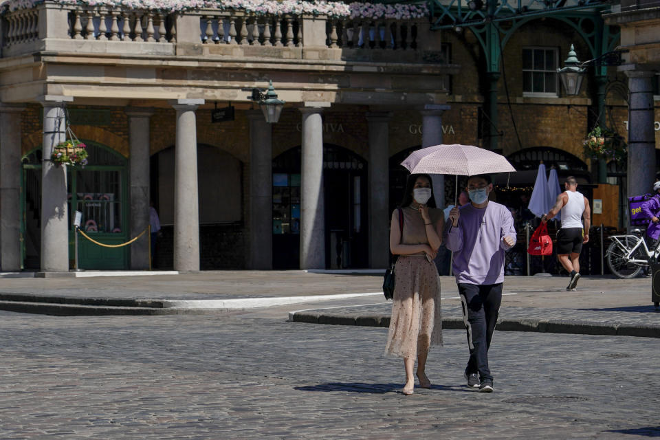 A couple wear face masks as they shelter from the sun under an umbrella, while they walk in Covent Garden, in London, Monday, June 14, 2021. British Prime Minister Boris Johnson is expected to confirm Monday that the next planned relaxation of coronavirus restrictions in England will be delayed as a result of the spread of the delta variant first identified in India. (AP Photo/Alberto Pezzali)