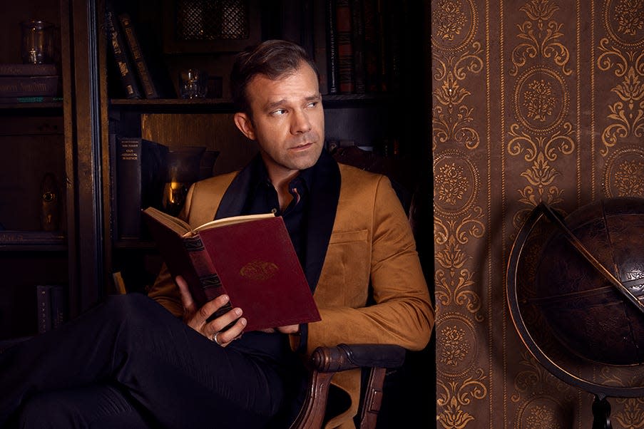 A wide shot of Critical Role main cast member Liam O'Brien. He's wearing a brown blazer and a black button-down shirt, and he's holding a red, leather-bound book.