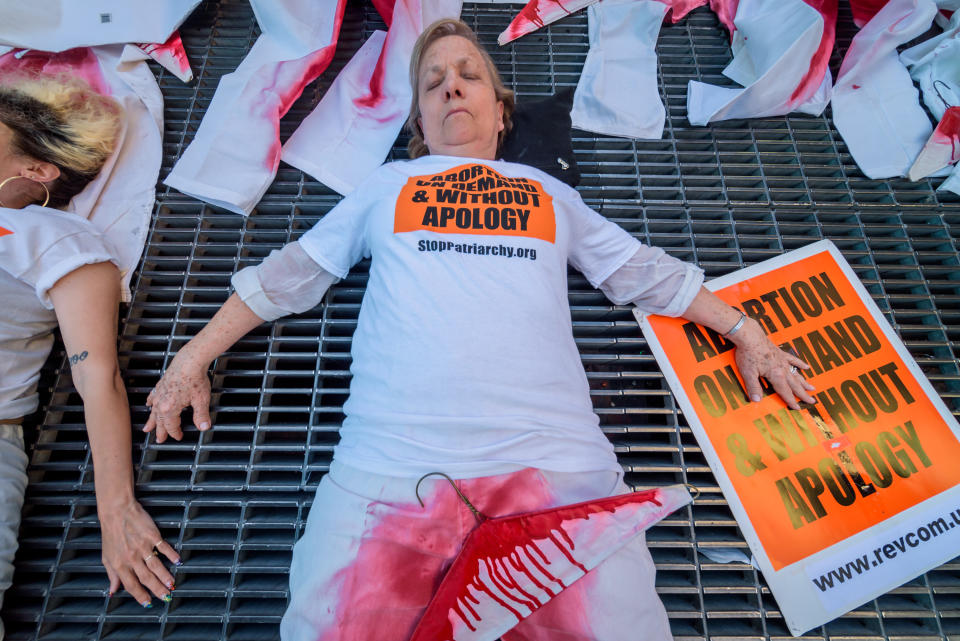 People lie on the ground with red-stained jeans and bloody coat hangers near signs reading "abortion on demand and without apology"