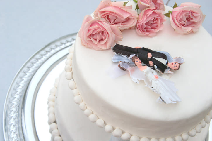 A wedding cake with pink roses on top featuring a broken bride-and-groom topper