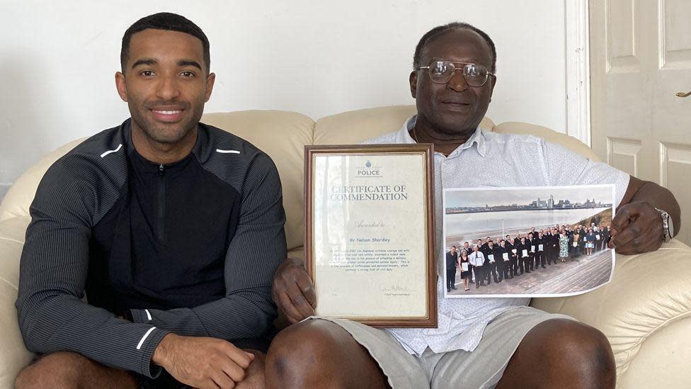 Nelson Shardey sits on his sofa holding his police commendation certificate, alongside his son Jacob