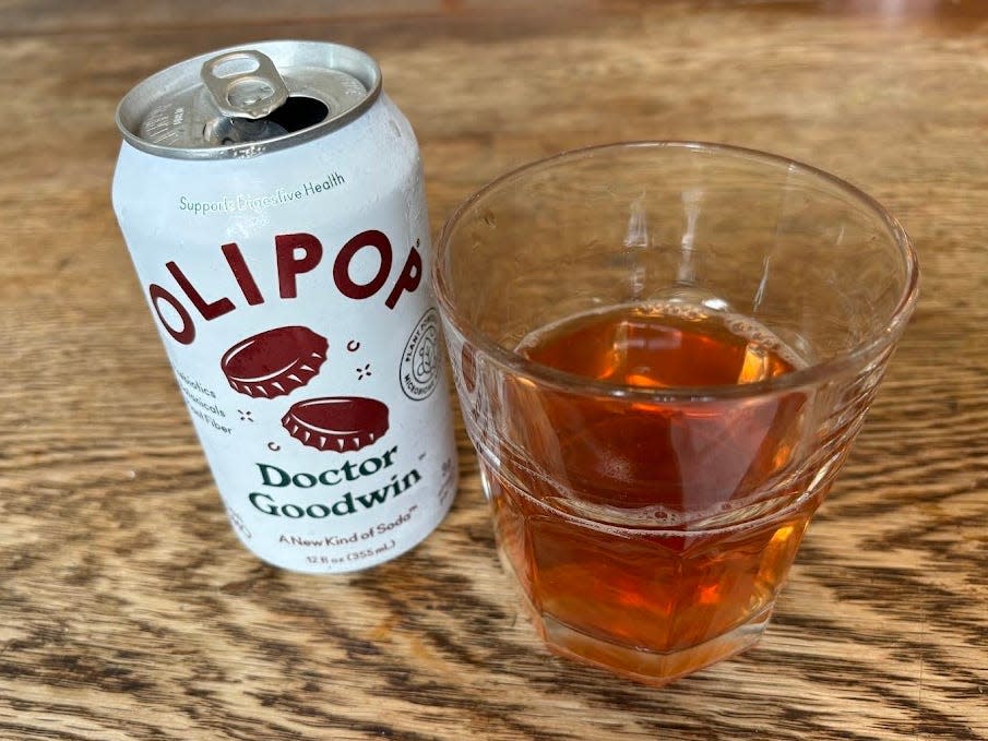 An open can of Doctor Goodwin Olipop next to a small, clear glass with brown liquid inside. Both are sitting on a wooden table.