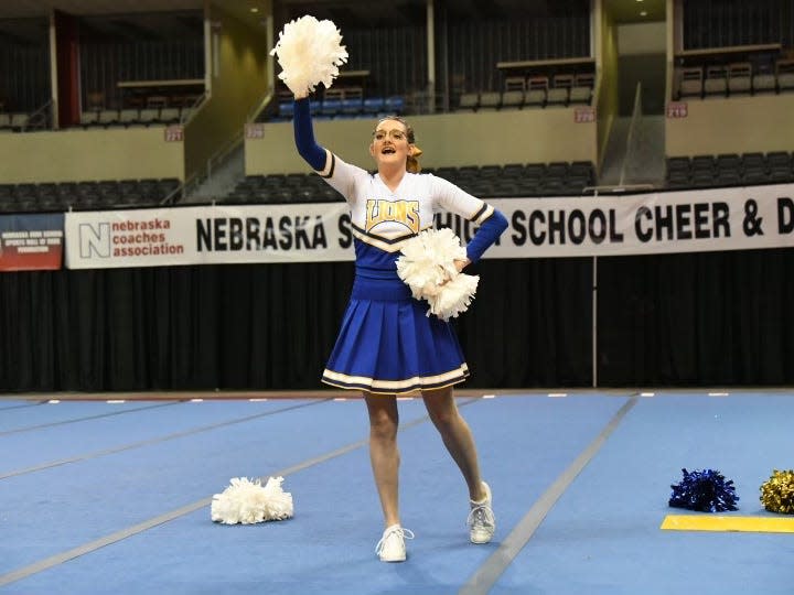 A cheerleader in a blue-and-white uniform holding white pom-poms raises her right arm. She's standing on a floor of blue mats, and a sign showing she's at a Nebraska high-school competition is in the background.
