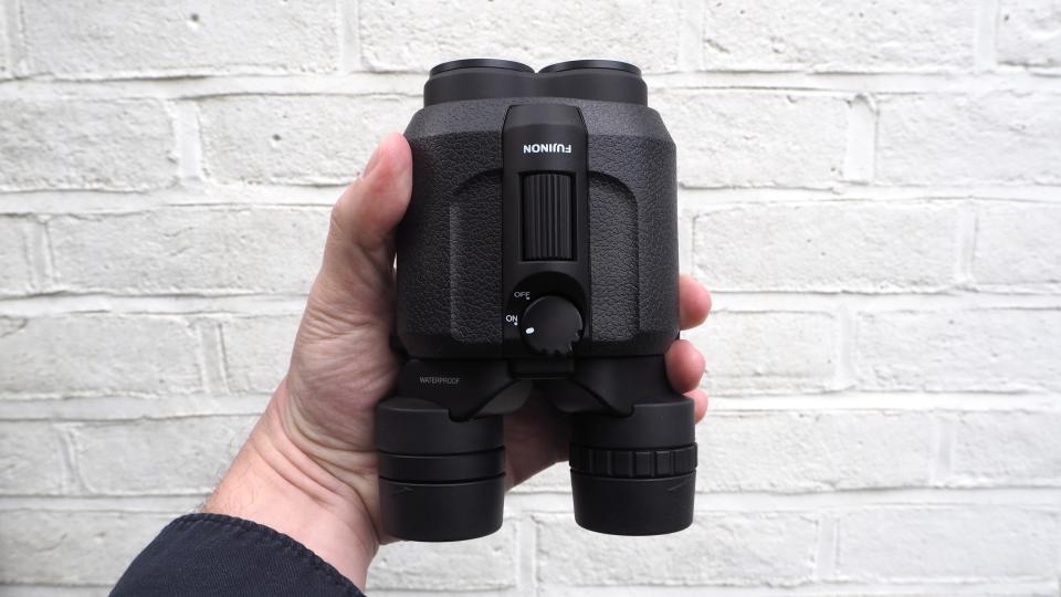 Fujifilm Fujinon Techno-Stabi TS16x28WP binocular on held in a hand in front of a white painted brick wall