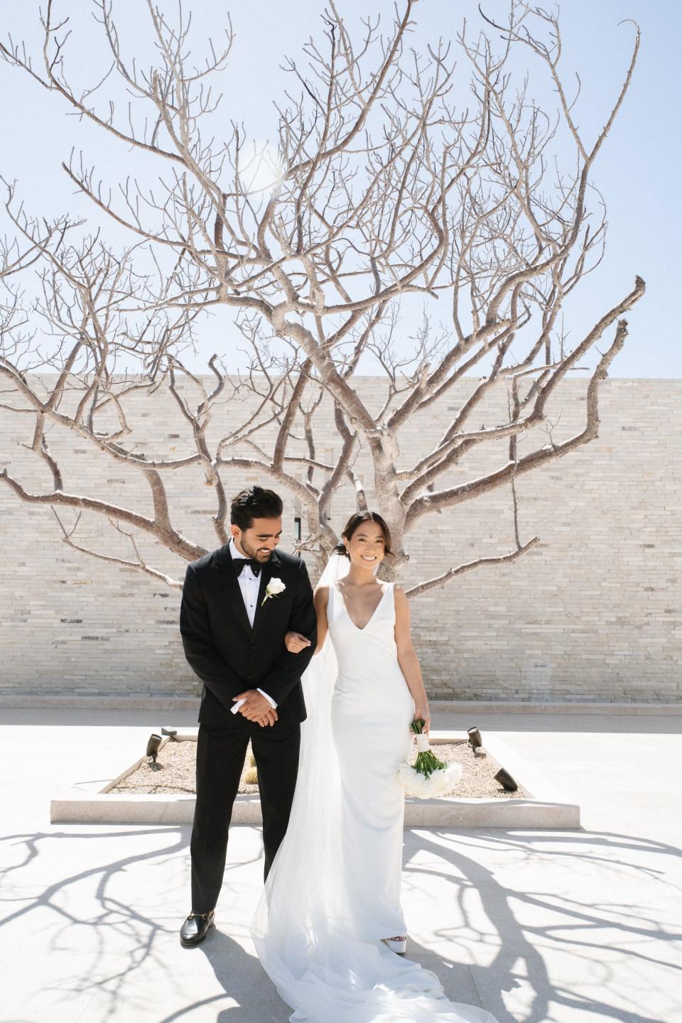 A groom looks at his bride in their wedding attire. They pose in front of a tree. 