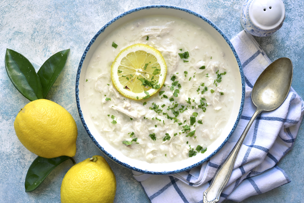 Top-view of Greek lemon and chicken soup, Avgolemono, in a blue-rimmed white bowl, a spoon on a blue and white checkered napkin on the right side, two lemons on the left side, on a light blue sponged tablecloth