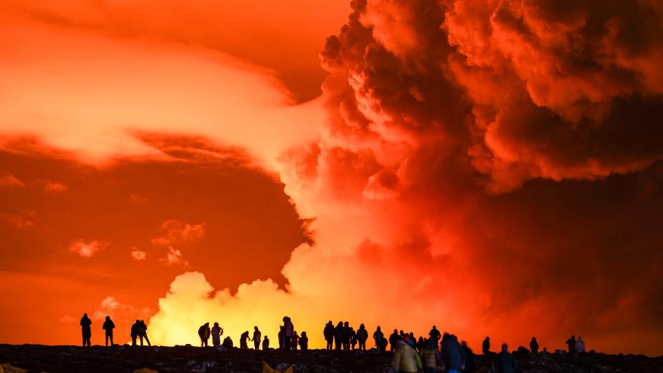 Eine große Gaswolke wird nachts von glühender Lava beleuchtet, während auftauchende Passanten zuschauen