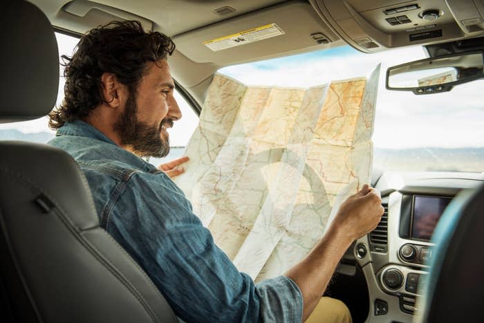 A man with wavy hair, wearing a denim jacket, sits in a car holding an unfolded map, looking at it intently, with mountains visible through the window