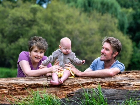 Nicci Attfield with her husband and their daughter Ava. Ava is a baby and sits on a log. Nicci and her husband sit behind her, holding Ava up.