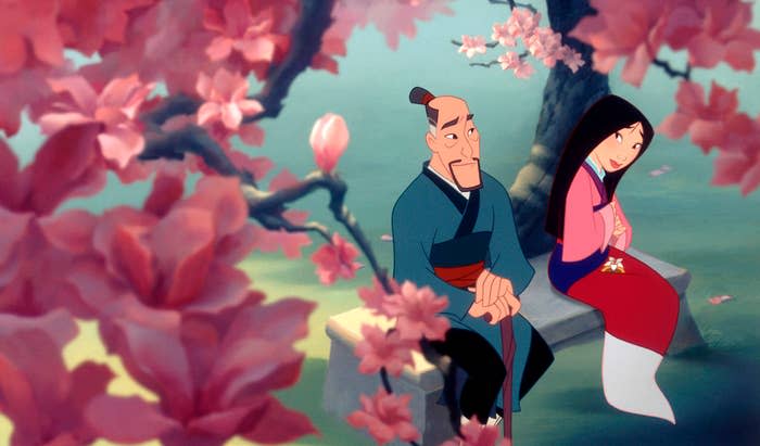 Fa Mulan and her father, Fa Zhou, sit on a bench under blooming cherry blossoms in a scene from the animated movie "Mulan."